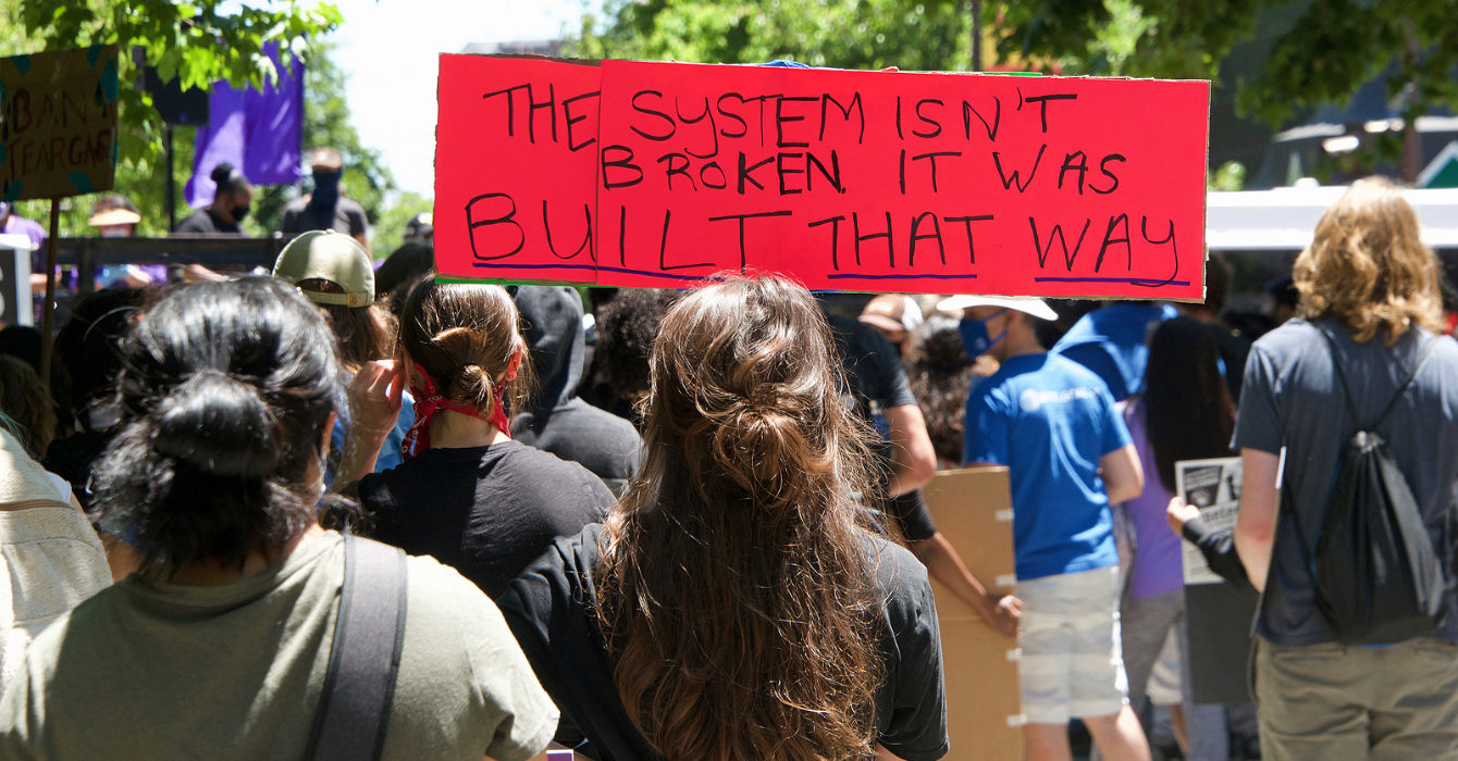 Protest sign reads "The system isn't broken it was built that way"