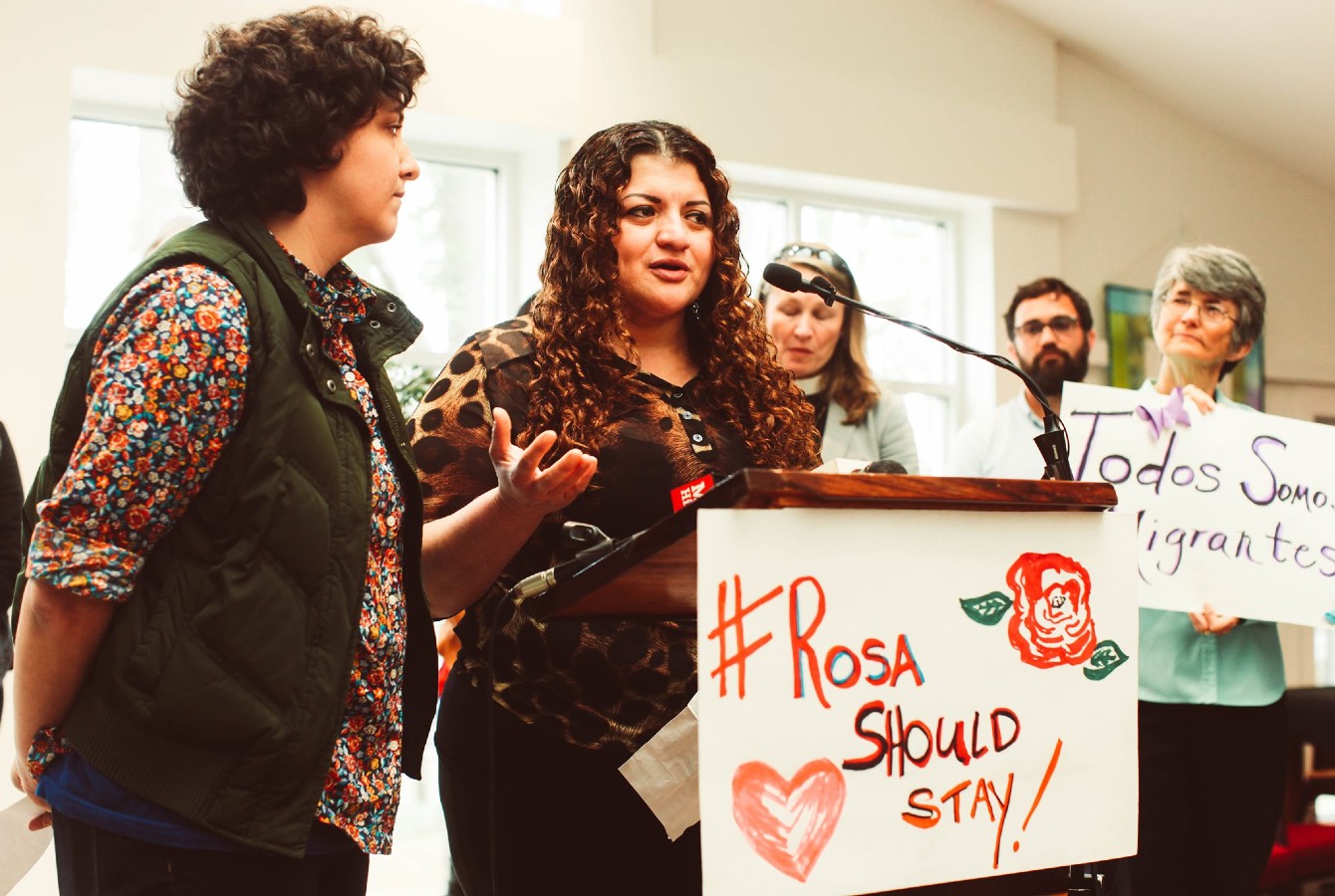 Image of a woman speaking at a podium with a sign that says "Rosa Should Stay"