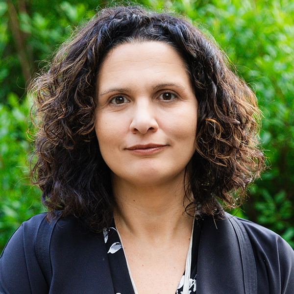 A woman with curly, shoulder-length hair looks into the camera. In the background are leafy, green trees.