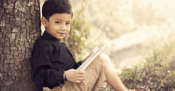 Boy sitting under a tree with a book looks at the viewer