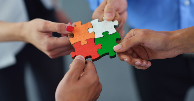Four people's hands, each holding a different jigsaw puzzle piece